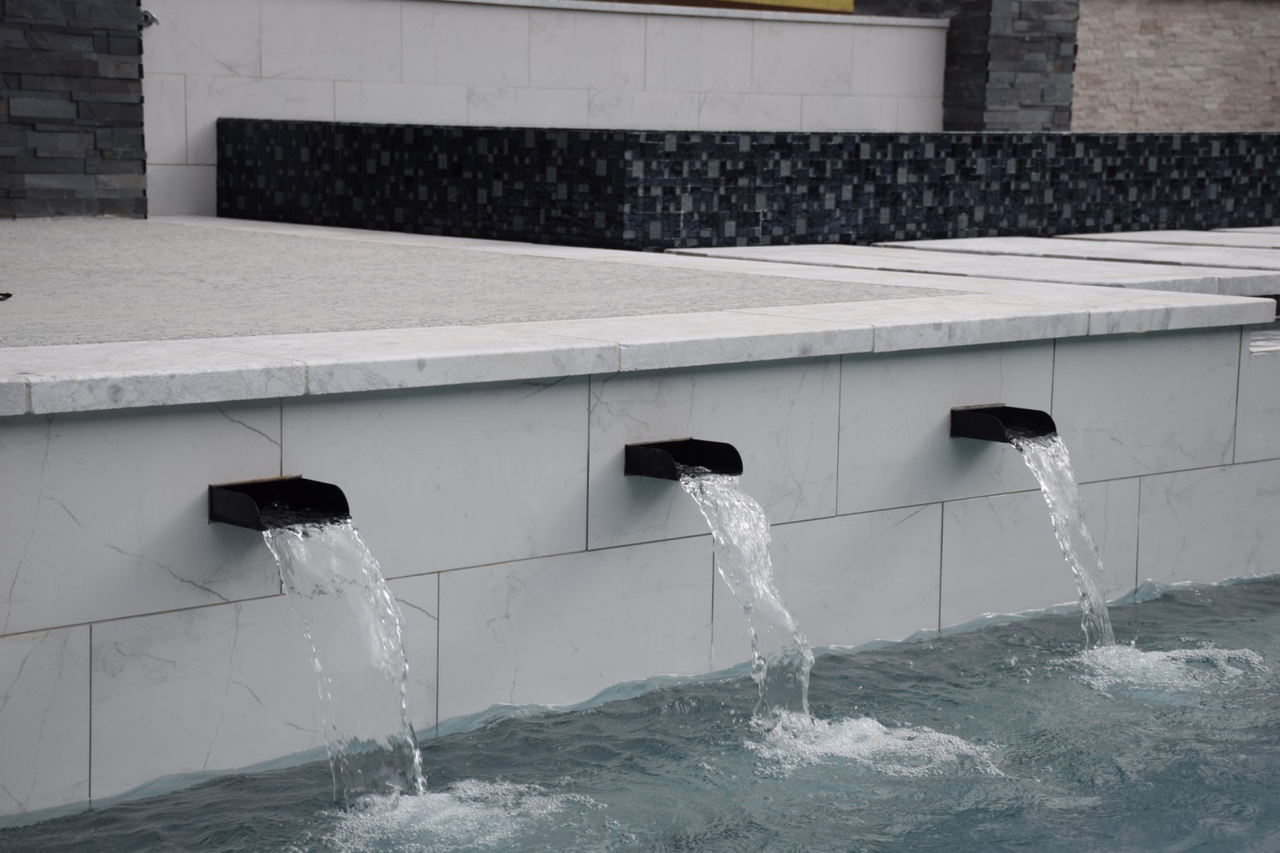 Three black scupper spouts pouring water into a renovated pool with new marble waterline tile