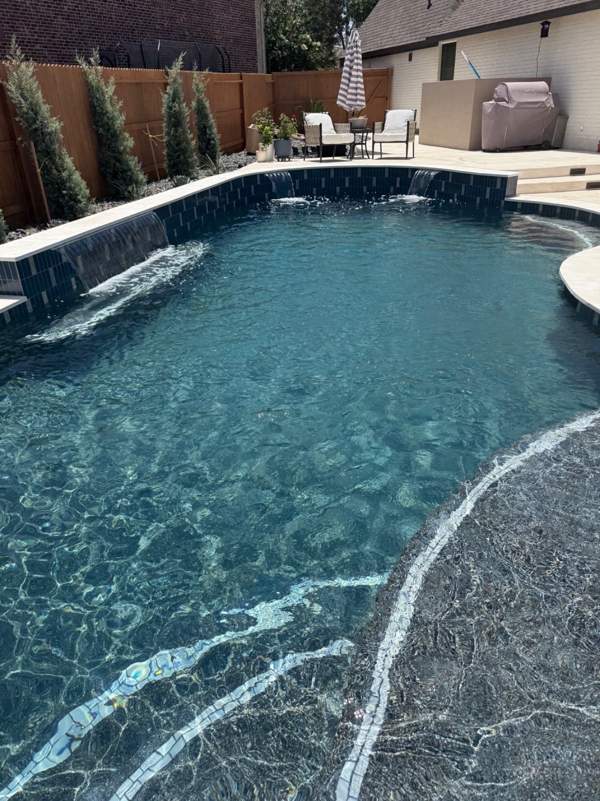 Freeform pool with twin sheer-descent water features spilling over a dark blue glass-tile wall, with a covered patio and outdoor kitchen beyond, in Veramendi, New Braunfels