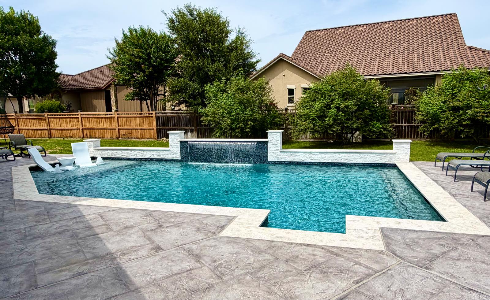 Geometric custom gunite pool with twin glass-tile sheer-descent water features, white stacked-stone wall, tanning ledge, and travertine deck built by J. Oliver Pools in New Braunfels