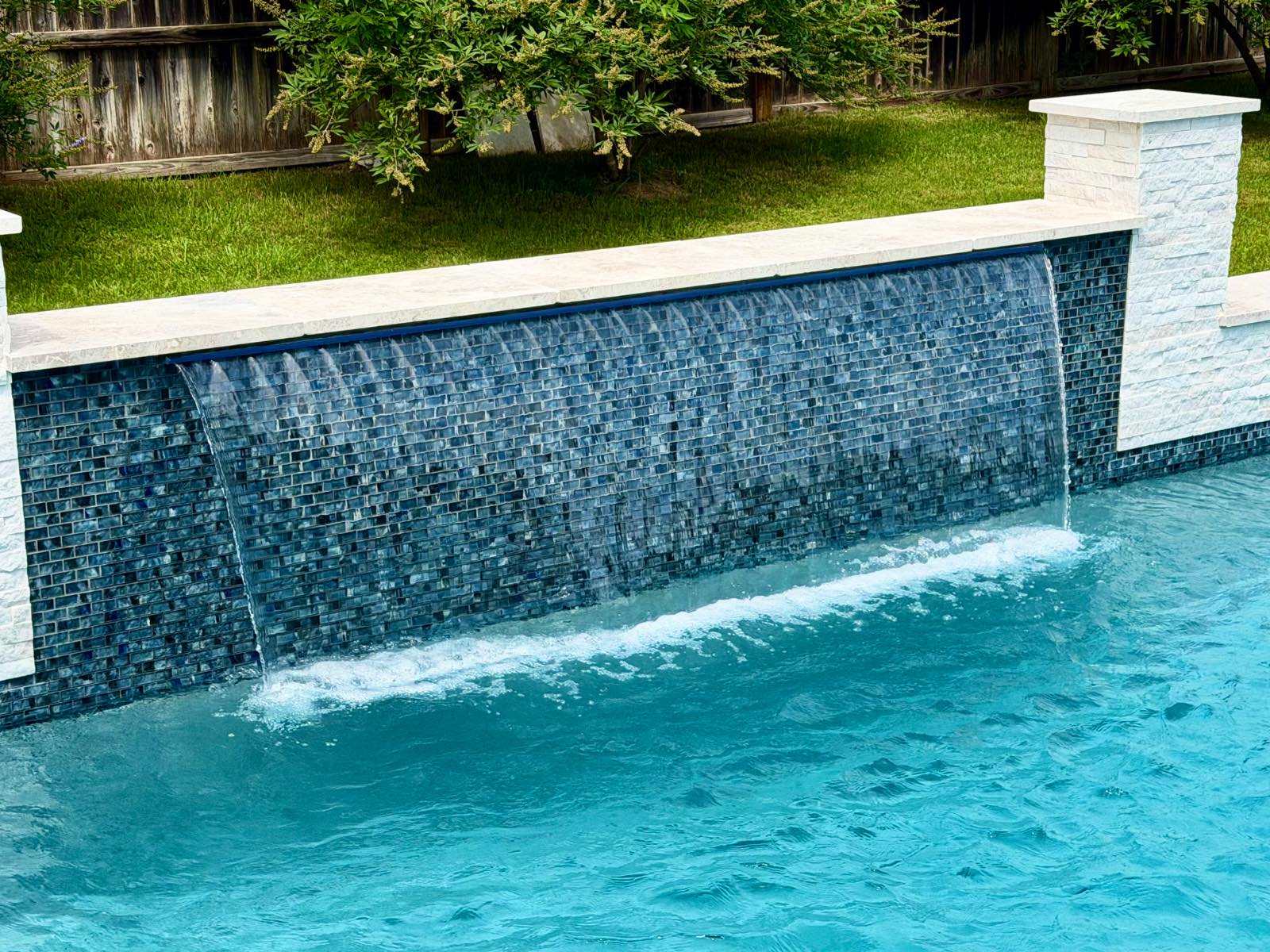 Close-up of a glass-tile sheer-descent waterfall spilling over a deep-blue mosaic wall capped with white limestone in New Braunfels