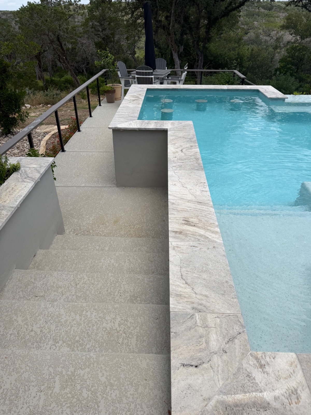 Raised bar-height sun shelf with travertine cap overlooking the vanishing-edge pool and Hill Country valley