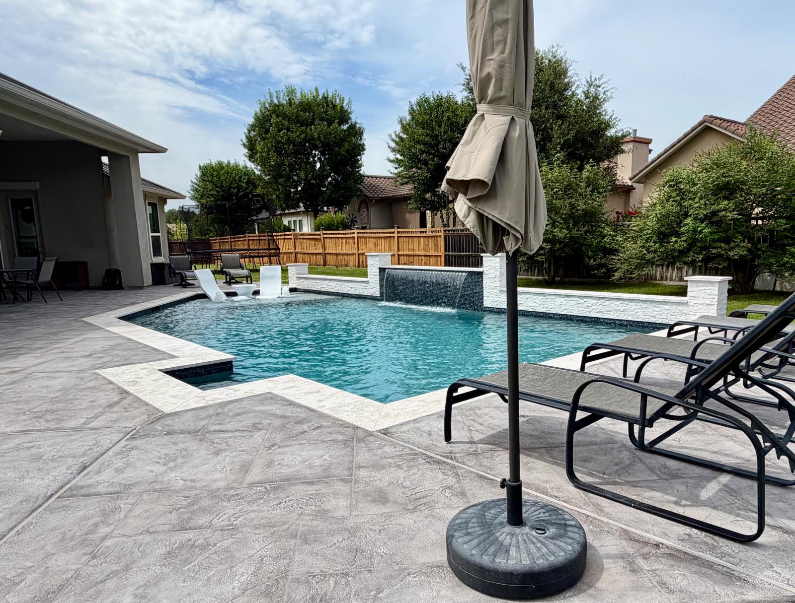 Stamped-concrete patio with loungers, shade umbrella, and travertine pool coping looking across the geometric pool toward the water wall in New Braunfels