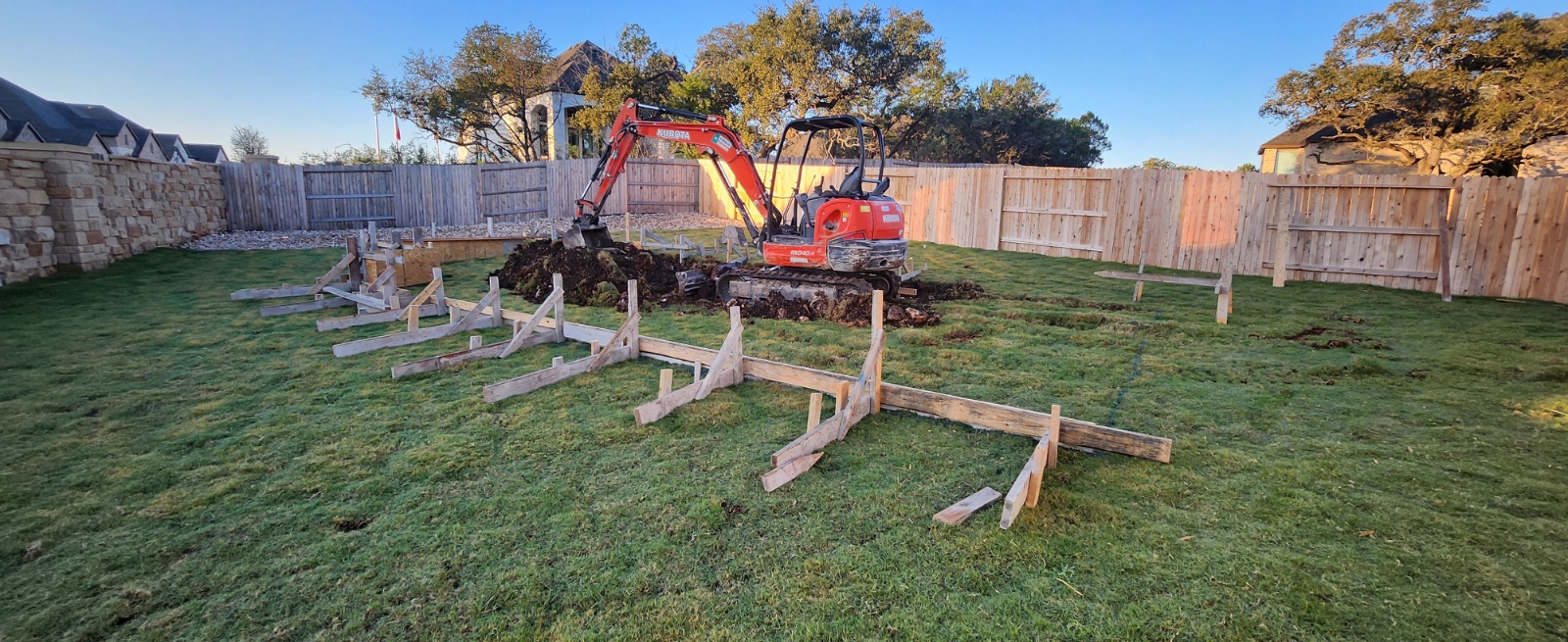 Pool layout staked out with wood forms across a grass new-build backyard in Veramendi, New Braunfels TX, with a mini excavator starting the dig