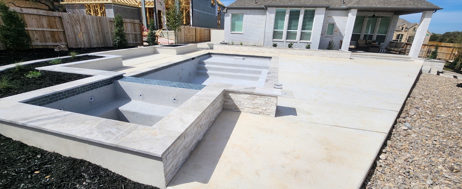 Finished French Grey PebbleTec pool with tanning ledge, bench seating, marble coping and raised spa, photographed before filling